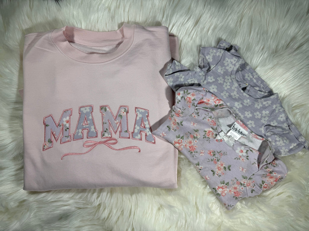Pink sweatshirt with 'MAMA' text and floral design, folded on a fluffy white surface.