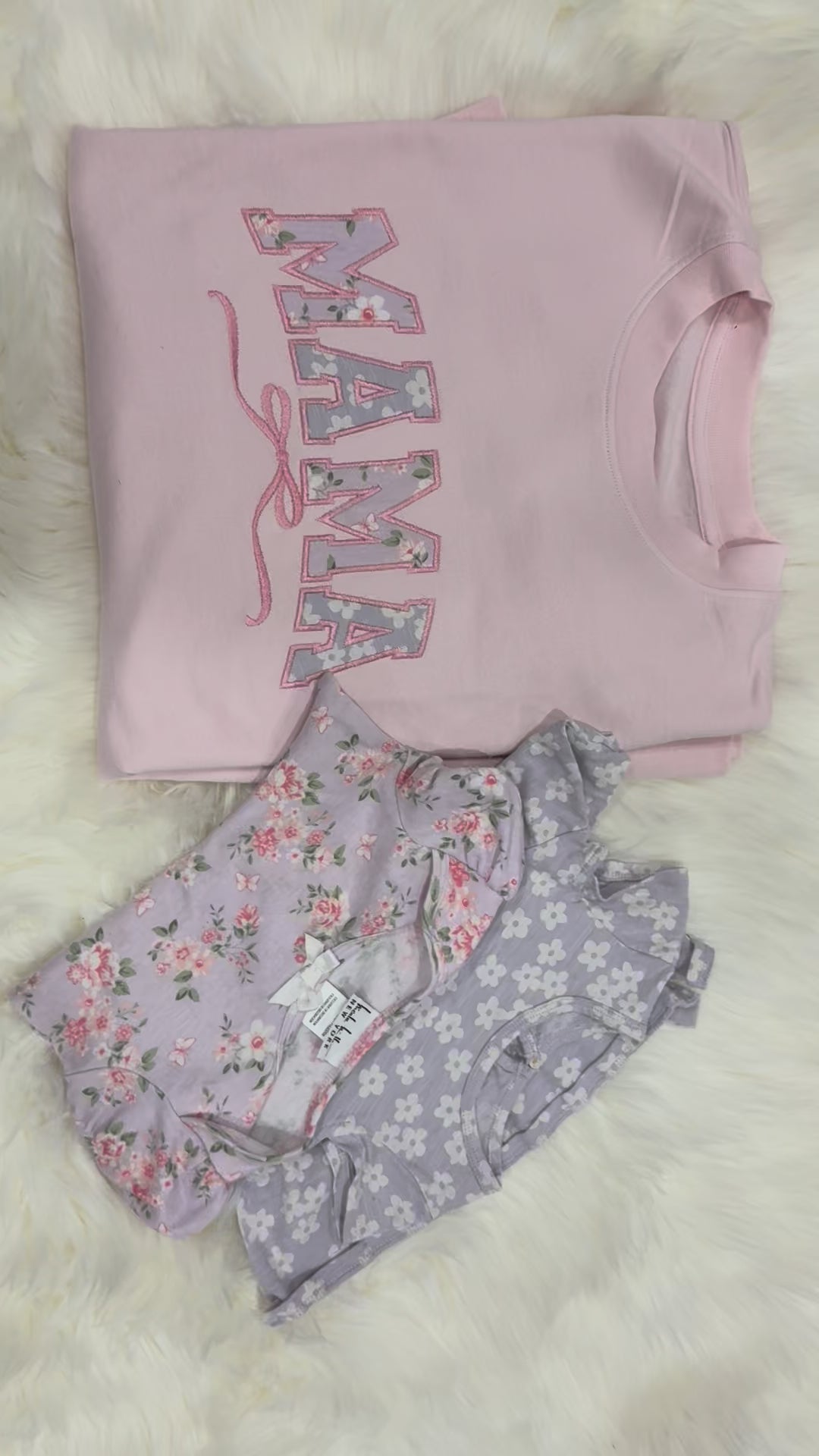 Pink 'MAMA' sweatshirt and floral baby outfit on a fluffy white surface